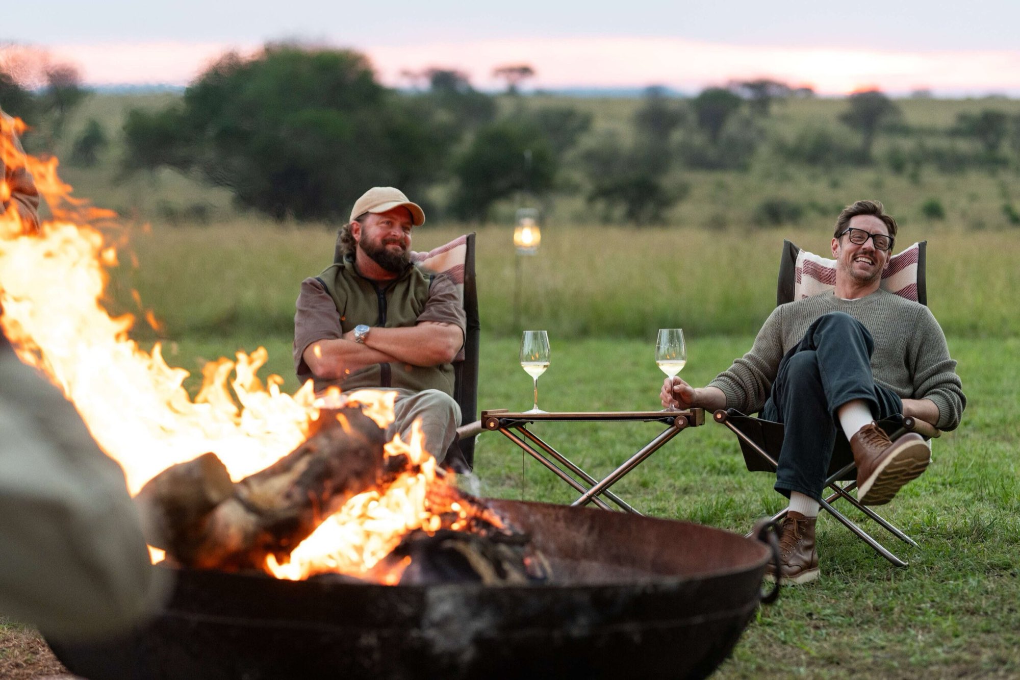 Two friends with sundowners beside a fire pit, looking out over the plains at dusk